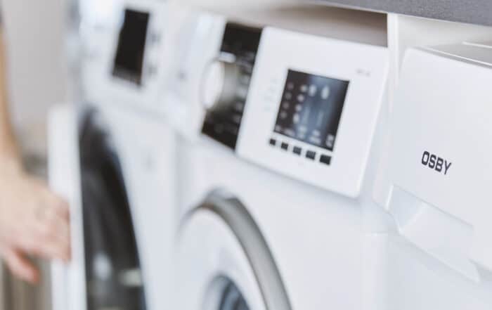 White front-loading washing machine with a digital control panel, in a blurred laundry-room setting