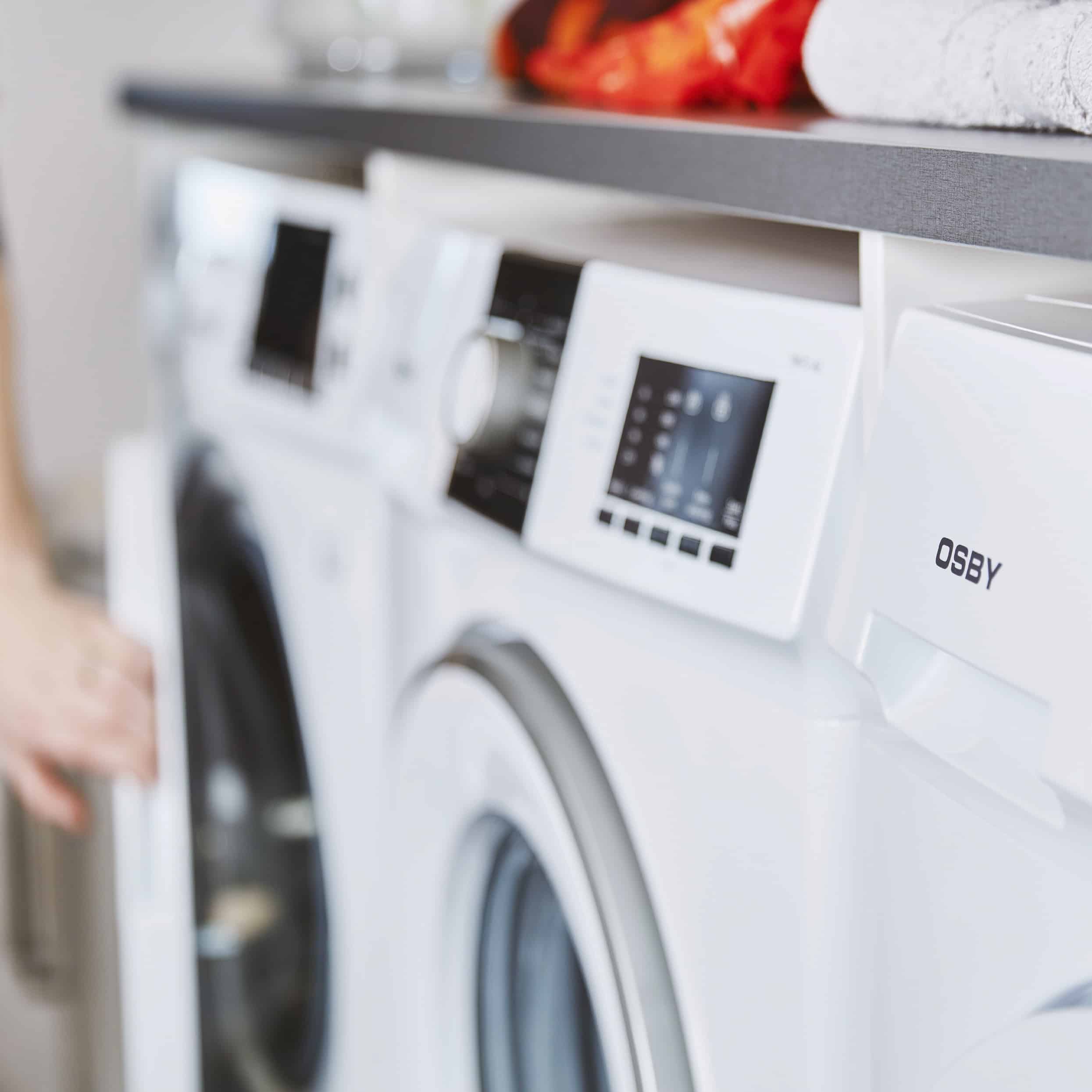 White front-loading washing machine with a digital control panel, in a blurred laundry-room setting